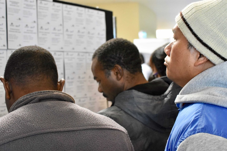 A group of men crowd around a board posted with job ads at the Sunshine Jobs Fair.