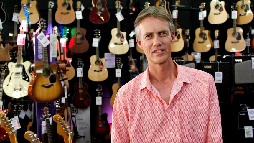 A man with short hair wearing a red and white shirt, standing in front of a wall covered in guitars.