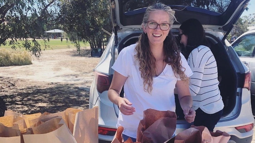 A lady in a white t-shirt stands by a table that is filled with bags of bread