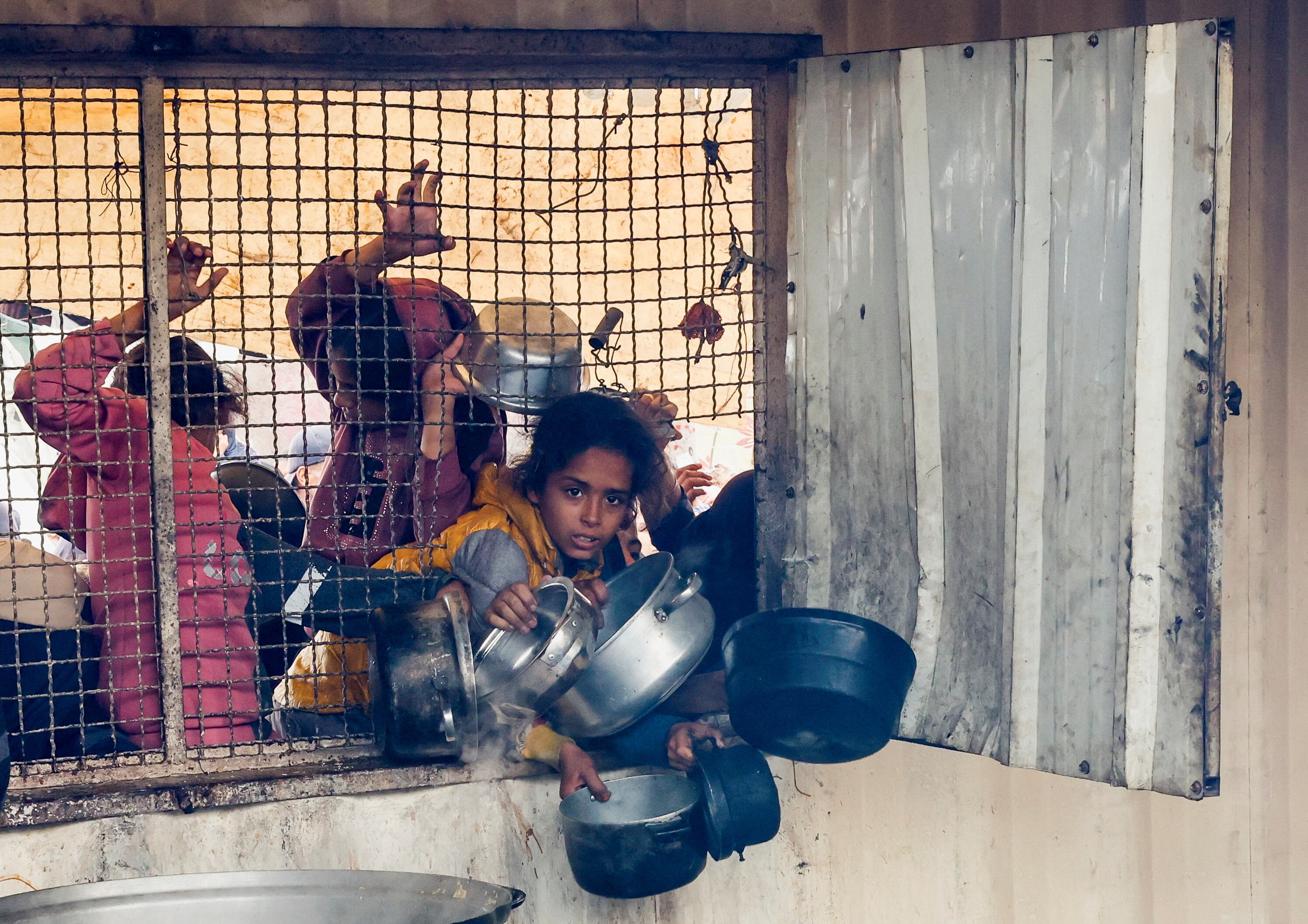 children hold bowls across a wire fence. 