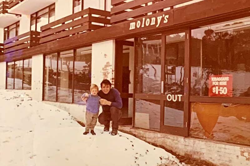 An old photo of a child and a man crouching in front of a ski shop, there is white writing that reads Molony's.