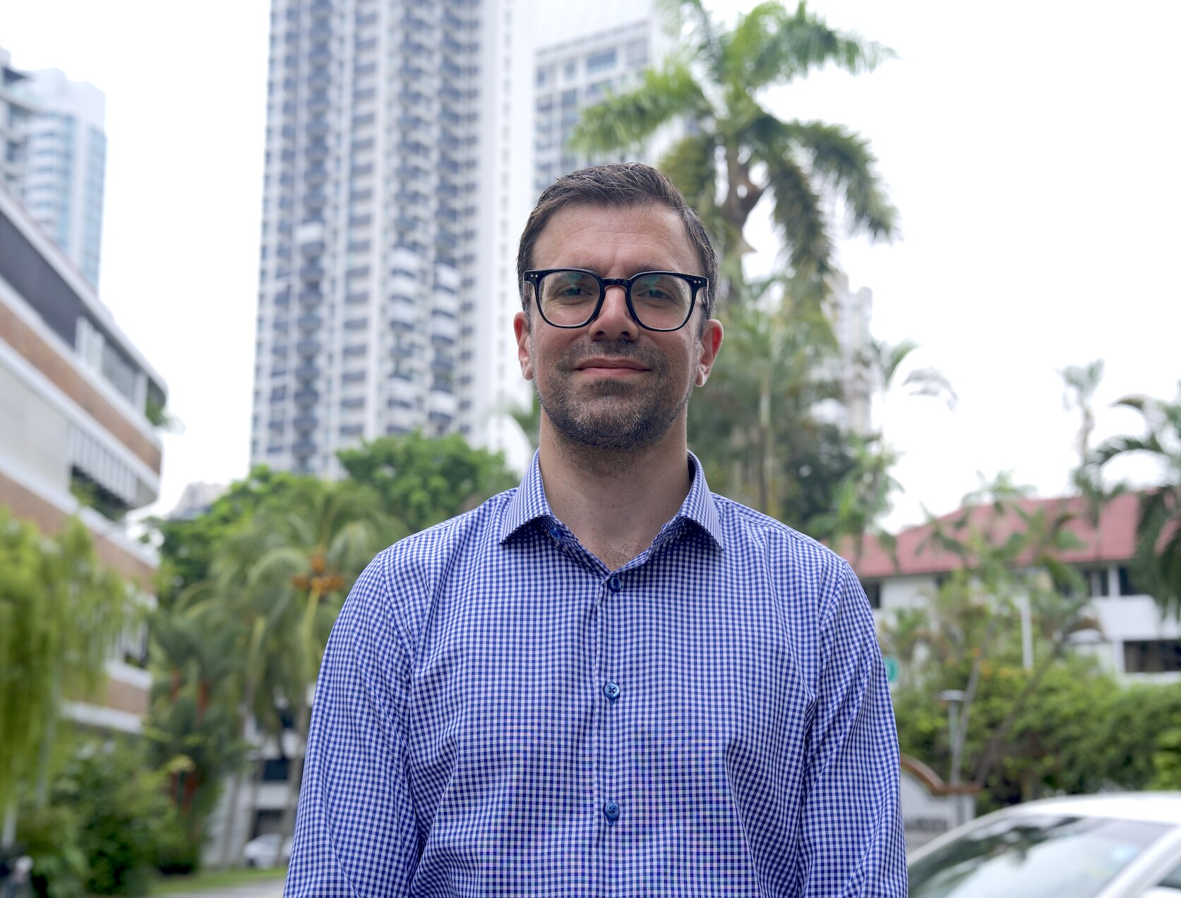 A bespectacled man in a button up top looks into the camera as he poses for a photo near high rises and palm trees.