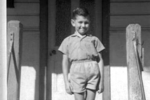 black and white image of young boy standing on front steps of house in school uniform