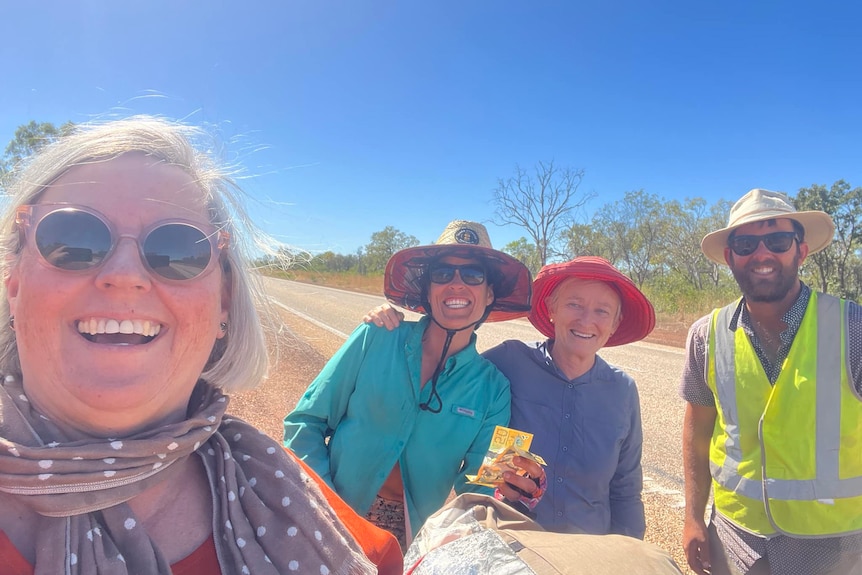 Three women and a man in hats and sun glasses smile for a selfie as a long road stretches out behind them.