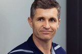 A smiling Justin Longmuir looks past the camera wearing a purple Fremantle Dockers shirt.