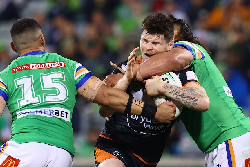 A Wests Tigers NRL player holds the ball while being tackled by two Canberra Raiders opponents.