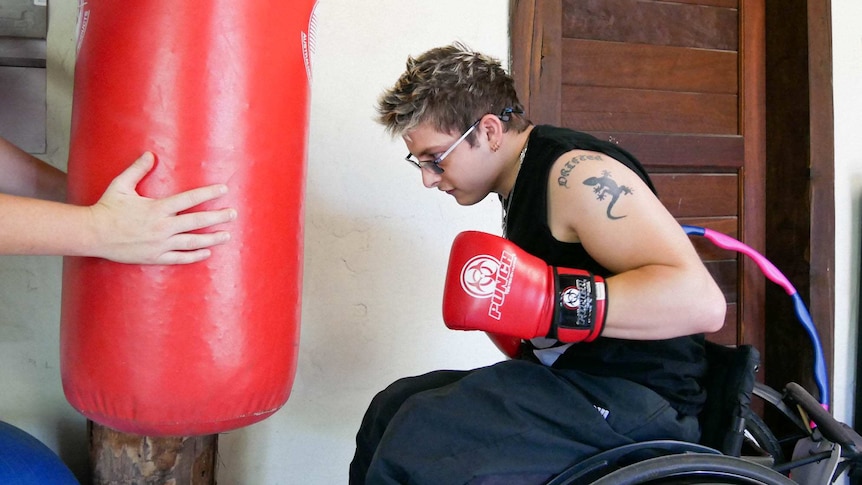 A man in a wheelchair and red boxing gloves on is about to hit a red punching bag being held by someone out of the picture.