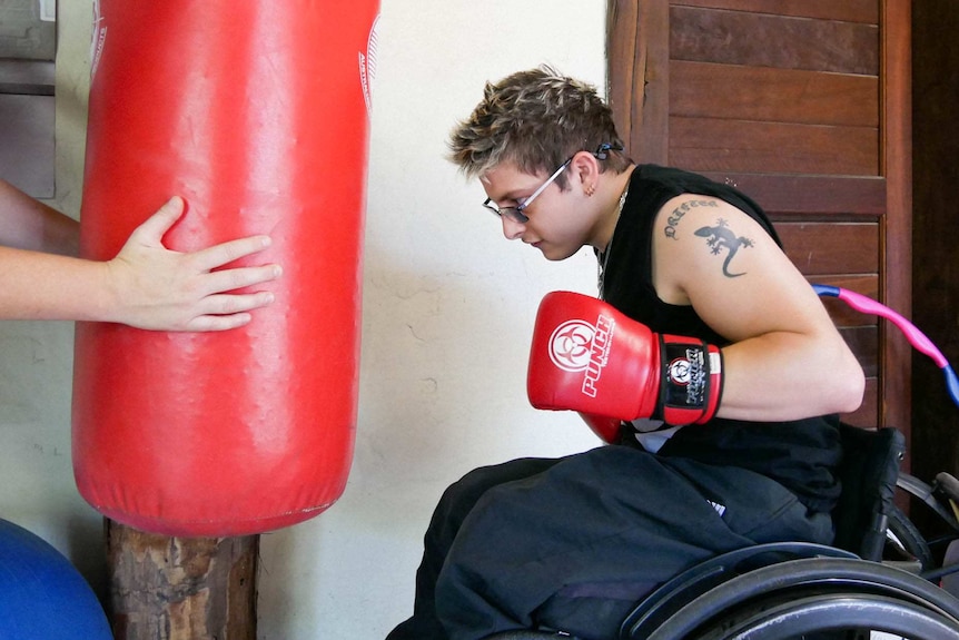 A man in a wheelchair and red boxing gloves on is about to hit a red punching bag being held by someone out of the picture.