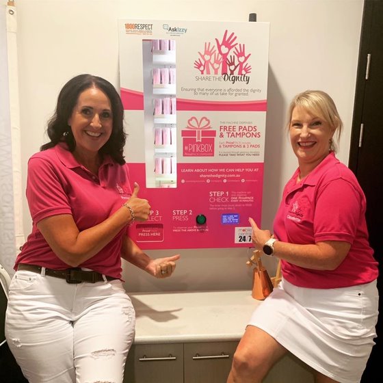 Two women wearing pink t-shirts smile in front of a period pack vending machine