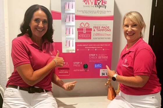 Two women wearing pink t-shirts smile in front of a period pack vending machine