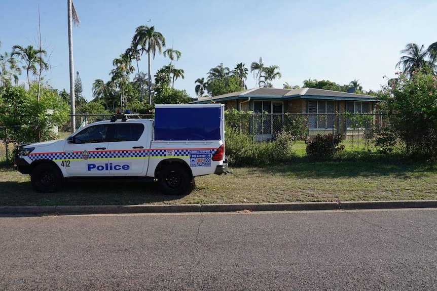 A police car is parked on the grassy verge, in front of a vine-covered metal fence surrounding a one story home.