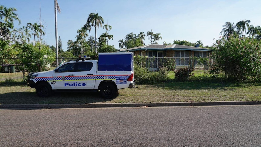 A police car is parked on the grassy verge, in front of a vine-covered metal fence surrounding a one story home.