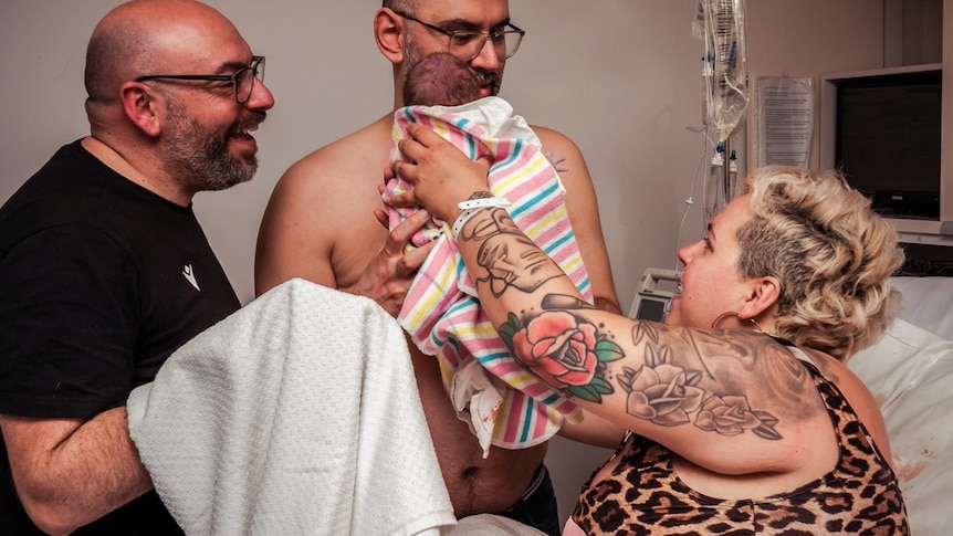 woman hands over a newborn baby to his two dads post birth
