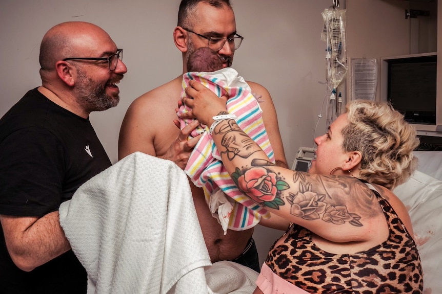 woman hands over a newborn baby to his two dads post birth