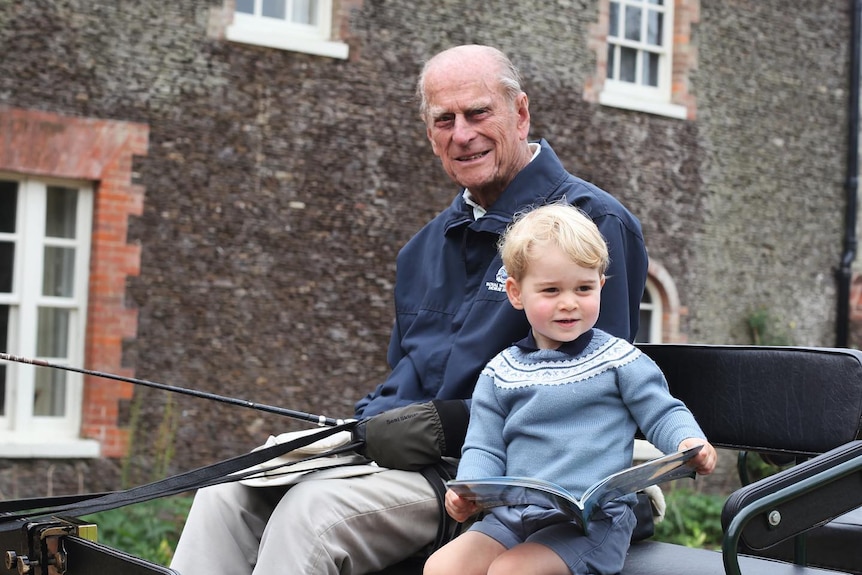 Princes Philip and Louis sit on the seat of an open top carriage. George holds open a picture book