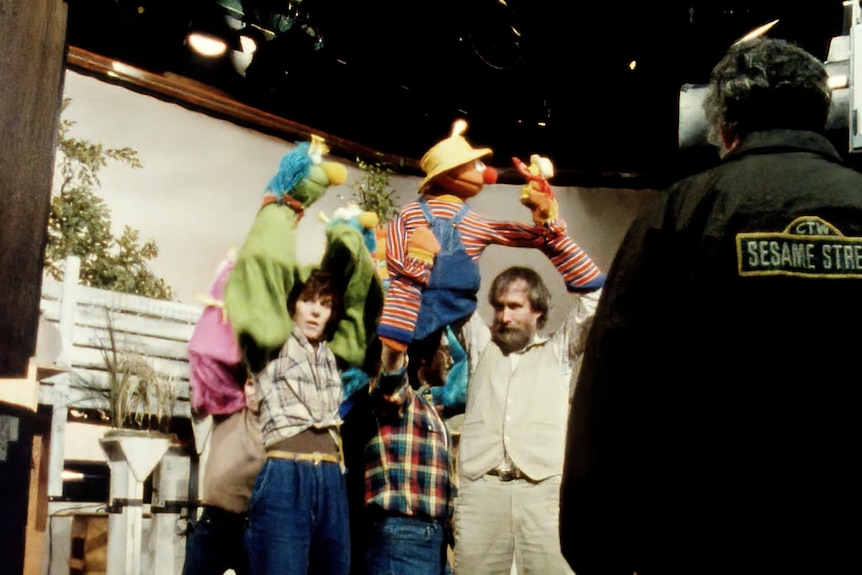 Three white men hold colourful puppets above their heads on a TV set while a camera man stands filming in the foreground.