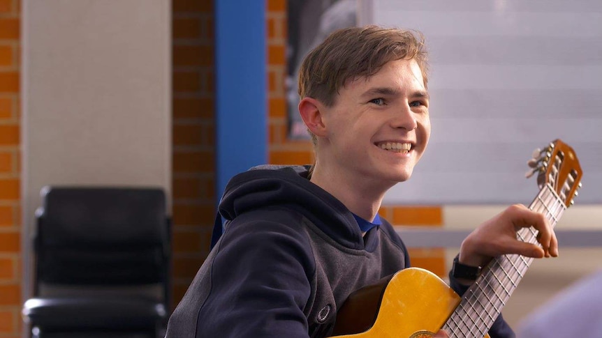 A young boy sits smiling, with his hand resting over the fretboard of a classical guitar.