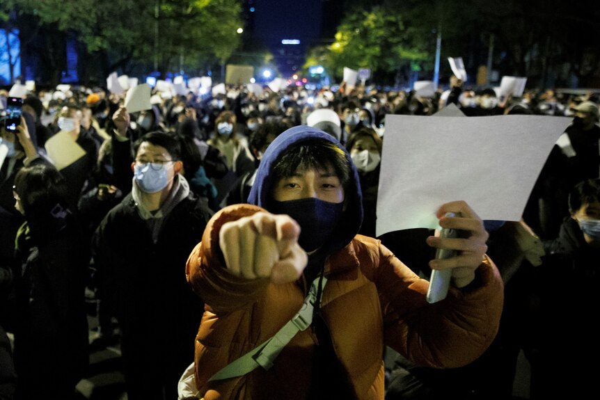 A large protest group, many with face masks and winter clothes, hold white sheets of paper, with one man pointing.