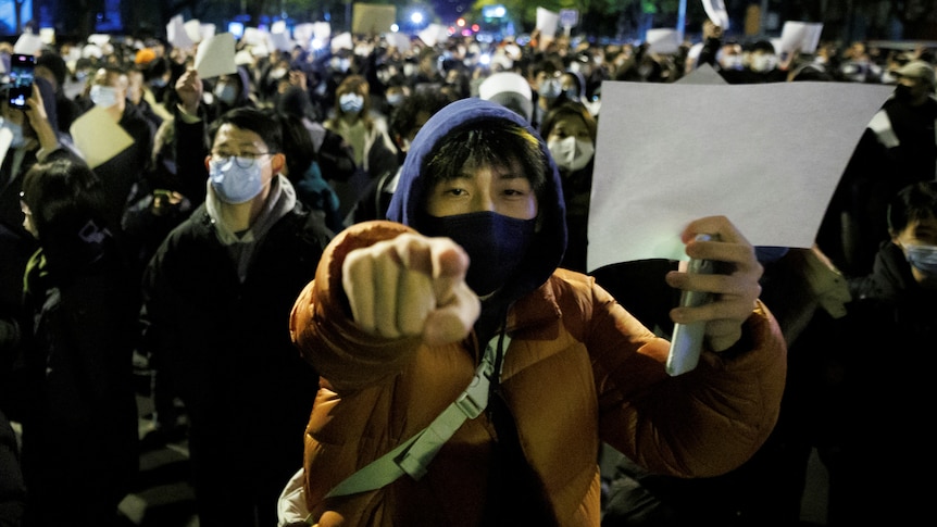 A large protest group, many with face masks and winter clothes, hold white sheets of paper, with one man pointing.