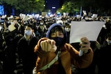 A large protest group, many with face masks and winter clothes, hold white sheets of paper, with one man pointing.