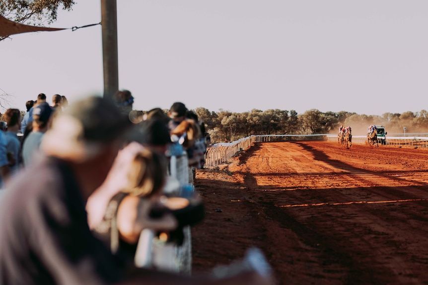 The crowd watches horses running at a red dirt country track. 