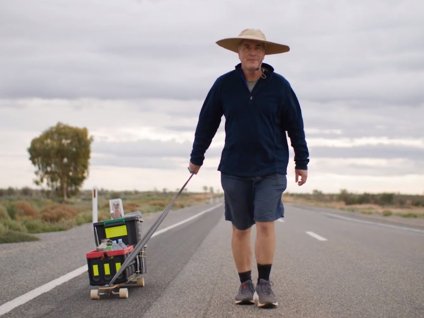 Dr Peter Hill walking with a wide brim hat along a road, pulling a skateboard carrying a crocheted fox on top near Broken Hill. 