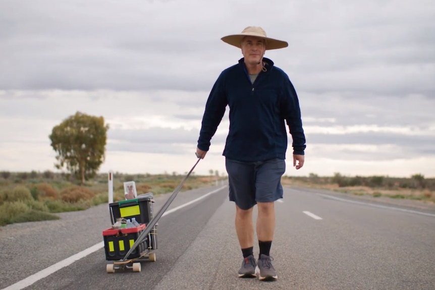 Dr Peter Hill walking with a wide brim hat along a road, pulling a skateboard carrying a crocheted fox on top near Broken Hill. 