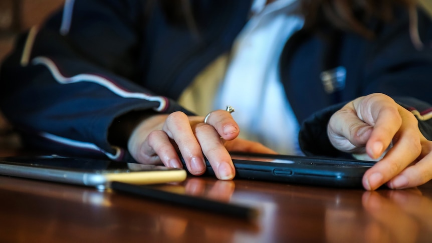 A generic image of a school student holding two phones on a desk strewn with pencils.