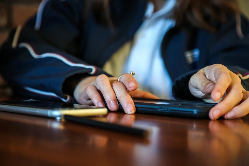 A generic image of a school student holding two phones on a desk strewn with pencils.