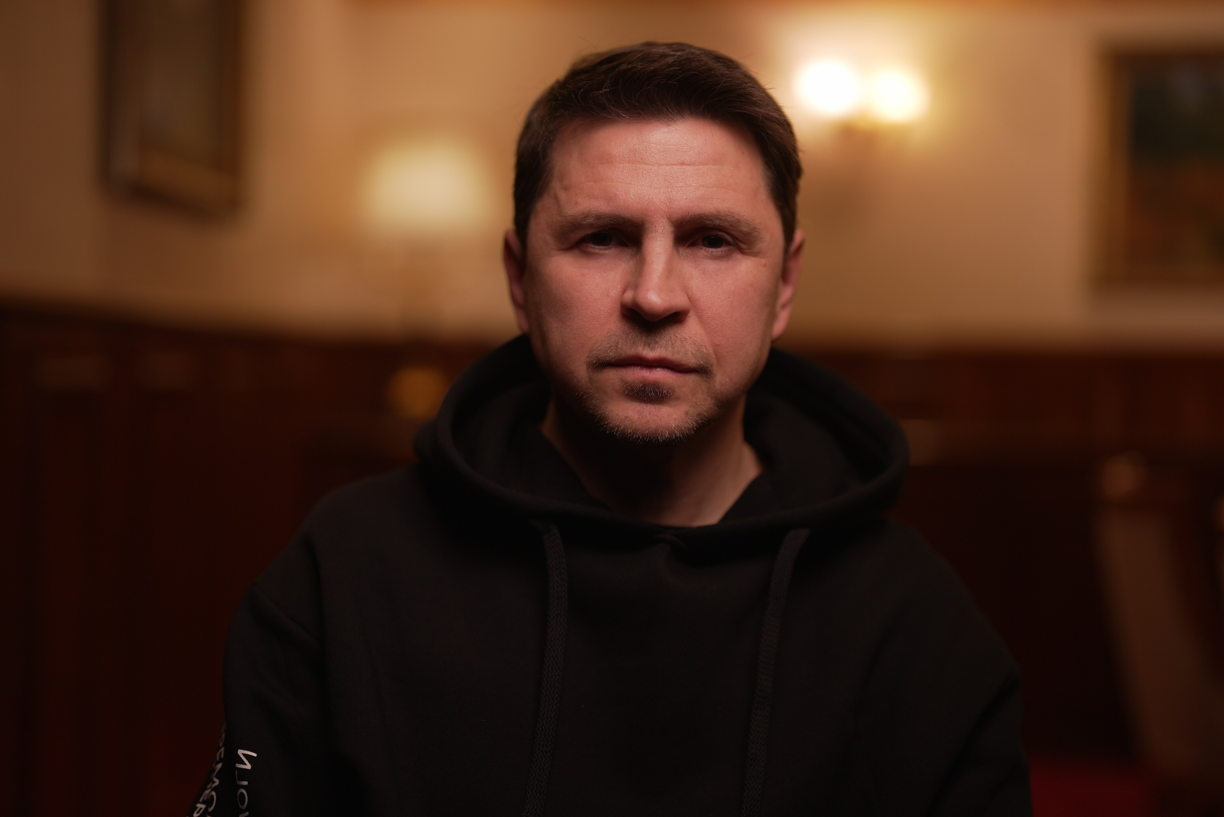 A man sitting indoors looks at the camera with a stern expression.