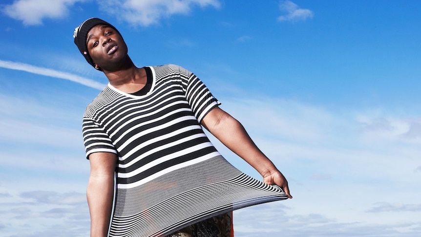 Cakes Da Killa stands in front of blue skies and clouds. They are wearing a black and white striped shirt and holding it out lik