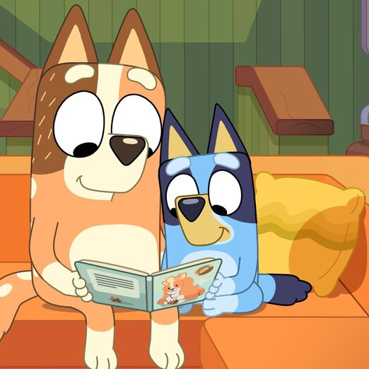 Still from Bluey showing two cartoon dogs sitting on the lounge reading a book.