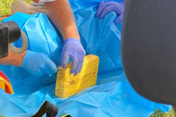 A close up of a body wrapped in tarp with a man wearing blue gloves hold a brick wrapped in yellow.