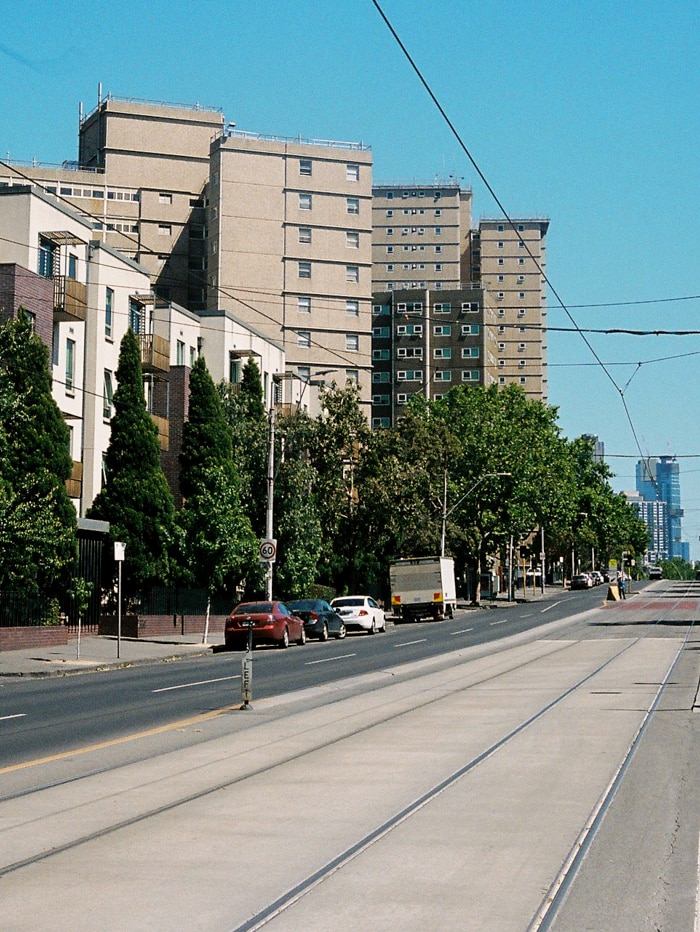 On a bright blue day, you see two multi-storey public housing towers behind newer townhouses off a road with tram tracks.