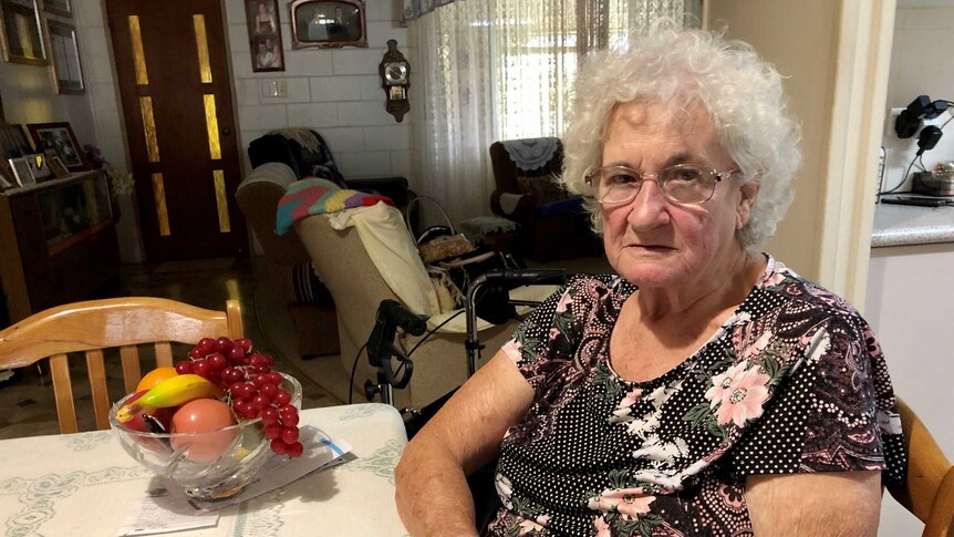 An elderly woman sits at her kitchen table with her mobility walker clearly visible in the background.