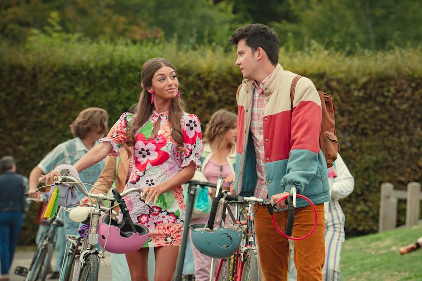 A teenage boy and girl, dressed in colourful clothes, walk their bikes as they chat outside school.