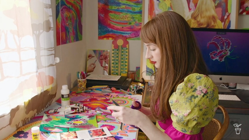 Woman wearing a colourful dress, sitting at a table cutting out brightly-coloured pictures.