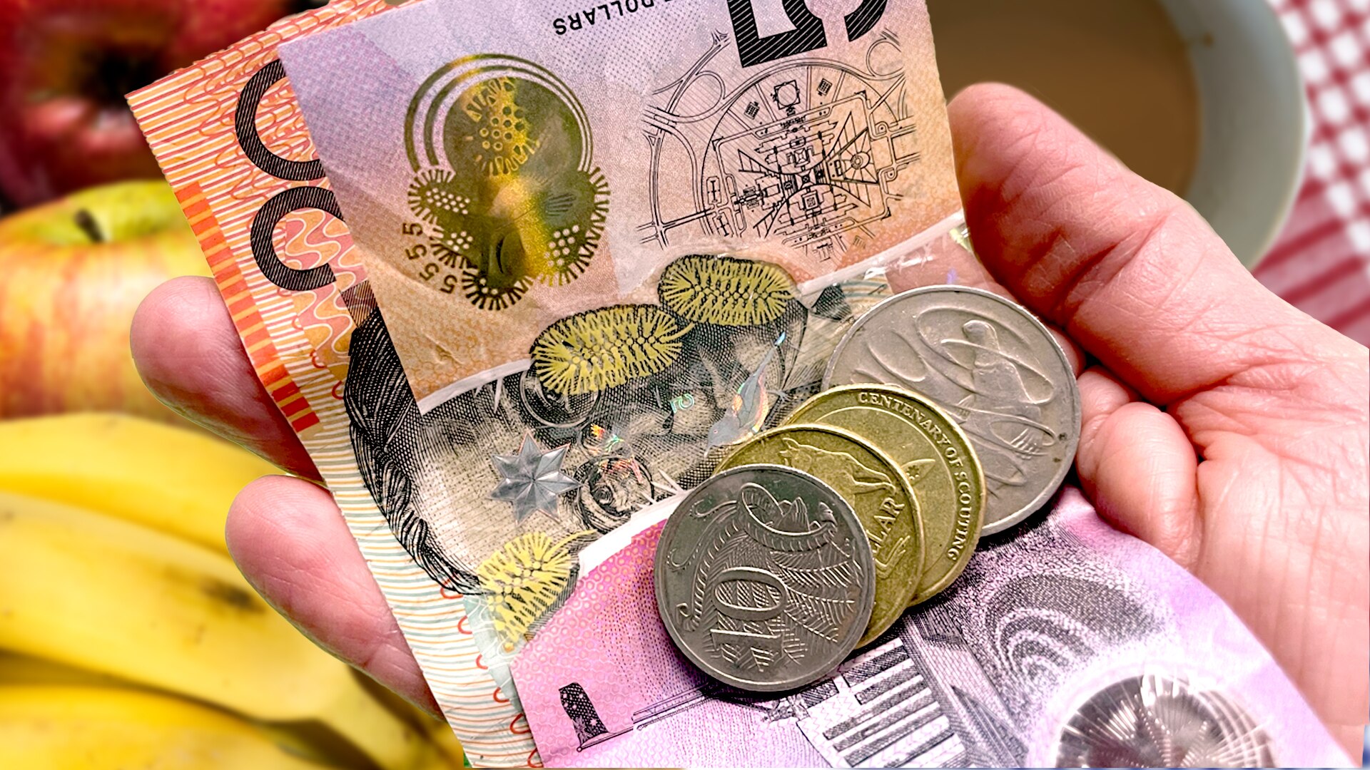 Hand holding money, notes and coins with a coffee cup and fruit in the background, cost of living