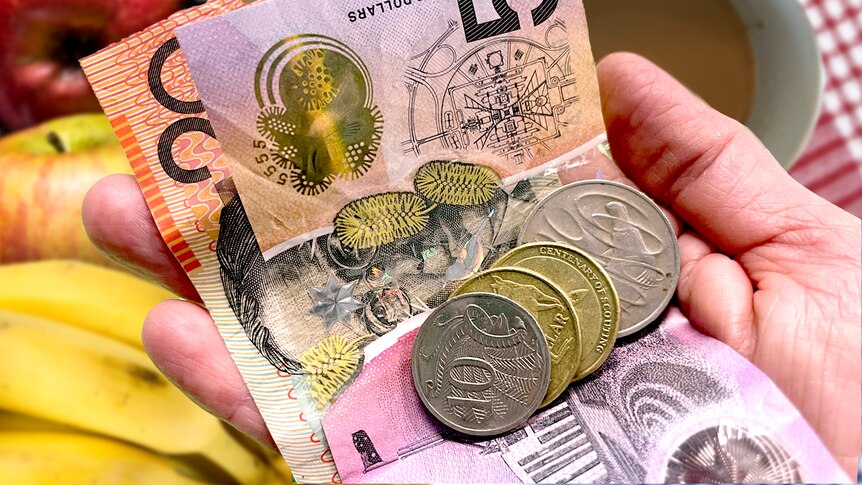 A hand holding a $20 and $5 note with coins in front of a banana and coffee mug