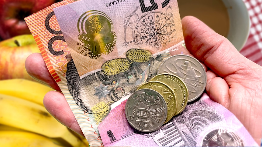 Hand holding money, notes and coins with a coffee cup and fruit in the background, cost of living