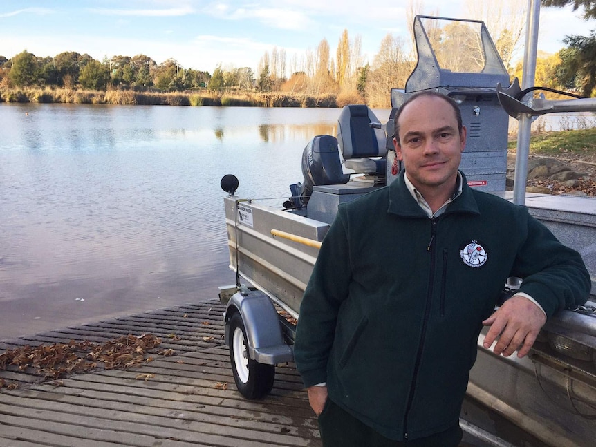 An ACT Government employee leans against a tin boat on the shores of a lake.