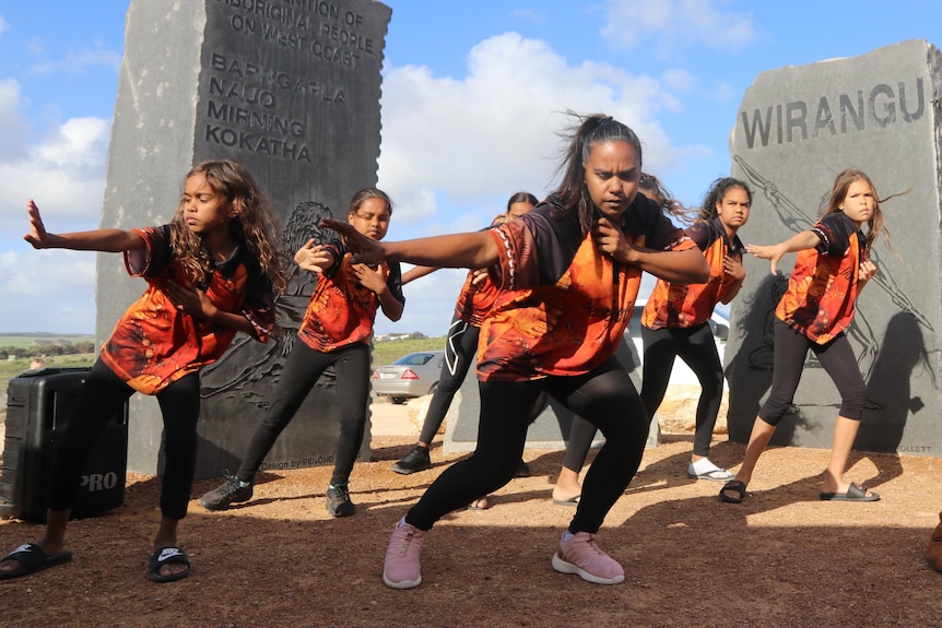 Girls in orange t-shirts dancing with arms reaching left in front of concrete memorial  