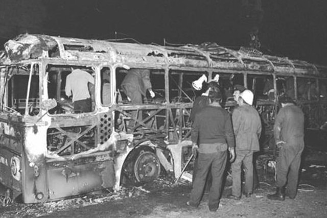 Several people walk through the remains of a burnt-out bus