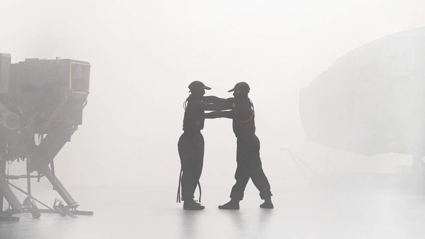 Hazy room, two dancers in black stand facing each other between two hulking machine forms, arms on each other's shoulders.