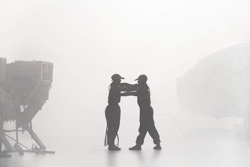 Hazy room, two dancers in black stand facing each other between two hulking machine forms, arms on each other's shoulders.