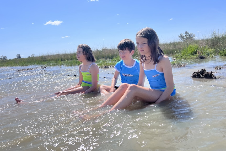 A girl, boy and another girl sit in their bathers, sit in a flooded paddock.