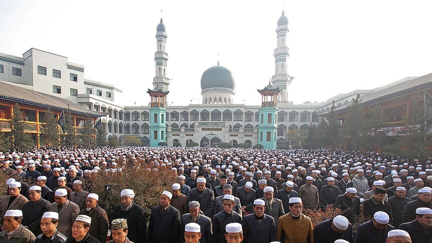 crowd of chinese muslims stand in front of larged domed mosque with minarets, wearing white caps on head