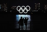 The five Olympic rings, are lit up and shining on the water in Tokyo as a couple watches from land.