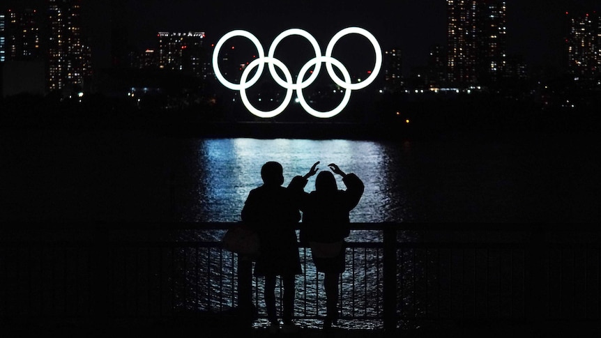 The five Olympic rings, are lit up and shining on the water in Tokyo as a couple watches from land.