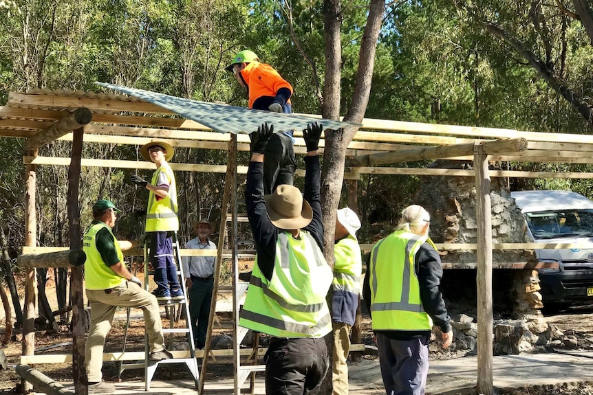 Workers in high-visibility clothes put a sheet of corrugated iron on a building's wooden frame.